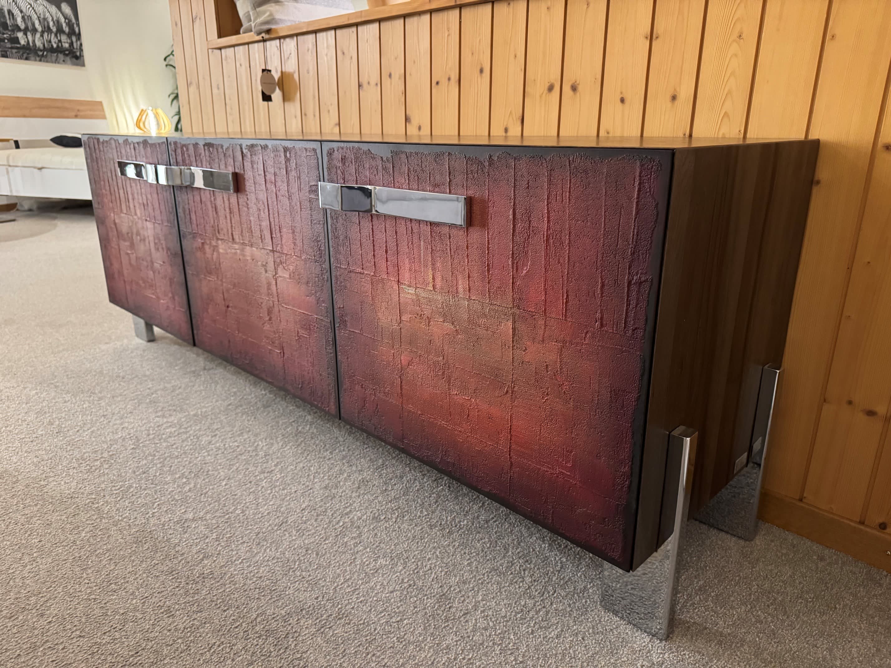 Das Sideboard Kredenz Gross hat rote Türen mit Struktur, Metallgriffe und -beine und wird vor einer holzgetäfelten Wand auf Teppichboden präsentiert. Das Sideboard Kredenz Gross hat rote Türen mit Struktur, Metallgriffe und -beine und wird vor einer holzgetäfelten Wand auf Teppichboden präsentiert.