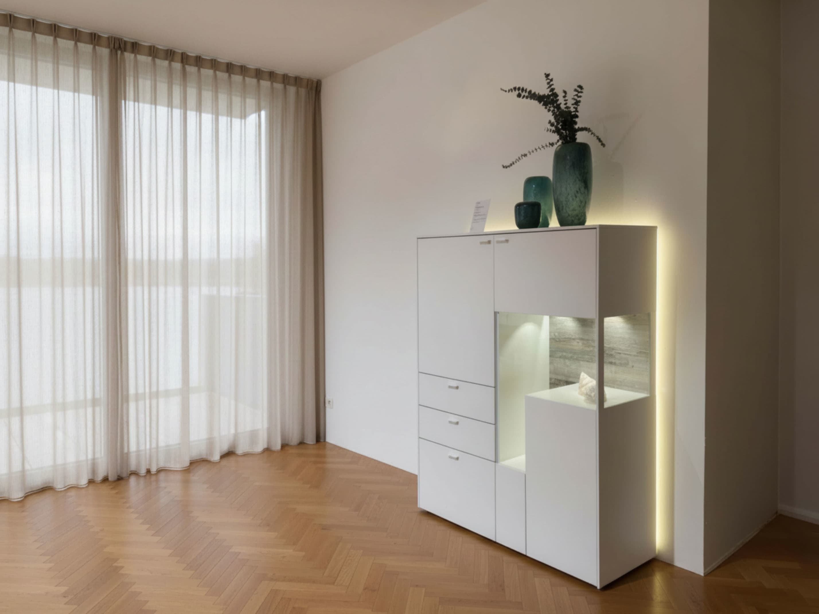 Minimalistisches Wohnzimmer mit transparenten Vorhängen, Holzfußboden und dem modernen Venjakob Highboard Sentino 3000 in Weiß, dekoriert mit stilvollen Vasen darauf. Minimalistisches Wohnzimmer mit transparenten Vorhängen, Holzfußboden und dem modernen Venjakob Highboard Sentino 3000 in Weiß, dekoriert mit stilvollen Vasen darauf.