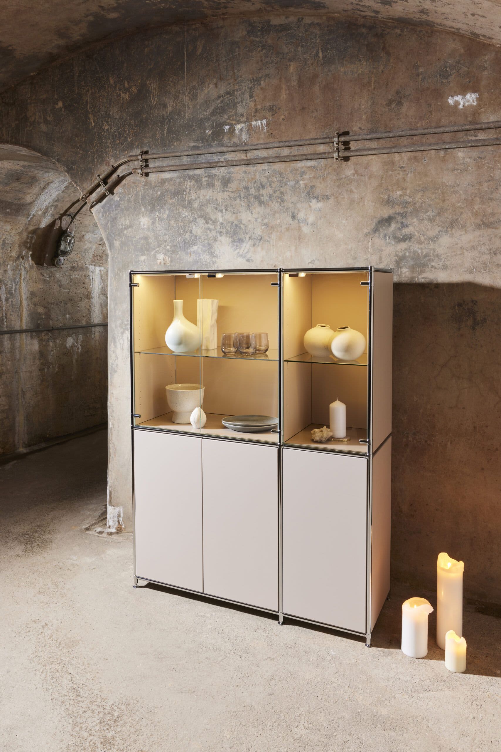 Die moderne Vitrine „MAB“ mit Keramikobjekten steht vor einer strukturierten Wand, flankiert von brennenden Kerzen auf dem Boden. Die moderne Vitrine „MAB“ mit Keramikobjekten steht vor einer strukturierten Wand, flankiert von brennenden Kerzen auf dem Boden.