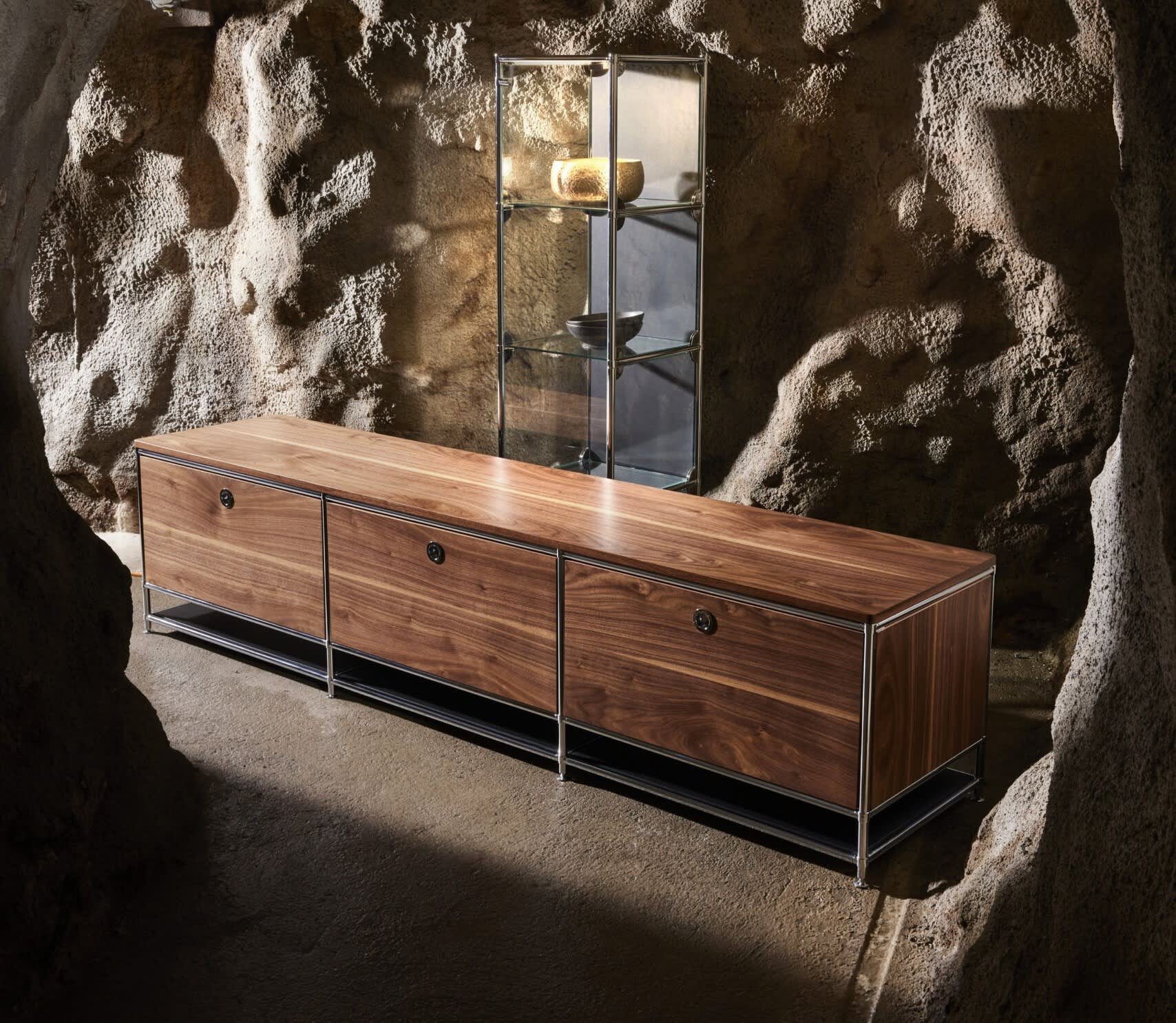 Das MAB-Sideboard „Celton“ mit seinem langgestreckten Holzdesign und Metallgestell steht in einer felsigen Höhle, im Hintergrund sind Glasregale und Töpferwaren zu sehen. Das MAB-Sideboard „Celton“ mit seinem langgestreckten Holzdesign und Metallgestell steht in einer felsigen Höhle, im Hintergrund sind Glasregale und Töpferwaren zu sehen.