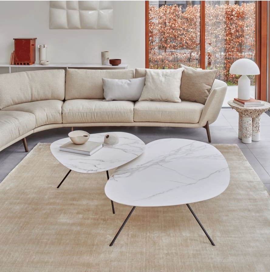 Un salon moderne avec un canapé beige et courbé, des tables d'appoint Leolux „Liliom“ en marbre, un tapis clair et de grandes fenêtres permettant de voir les arbres d'automne à l'extérieur. Un salon moderne avec un canapé beige et courbé, des tables d'appoint Leolux „Liliom“ en marbre, un tapis clair et de grandes fenêtres permettant de voir les arbres d'automne à l'extérieur.