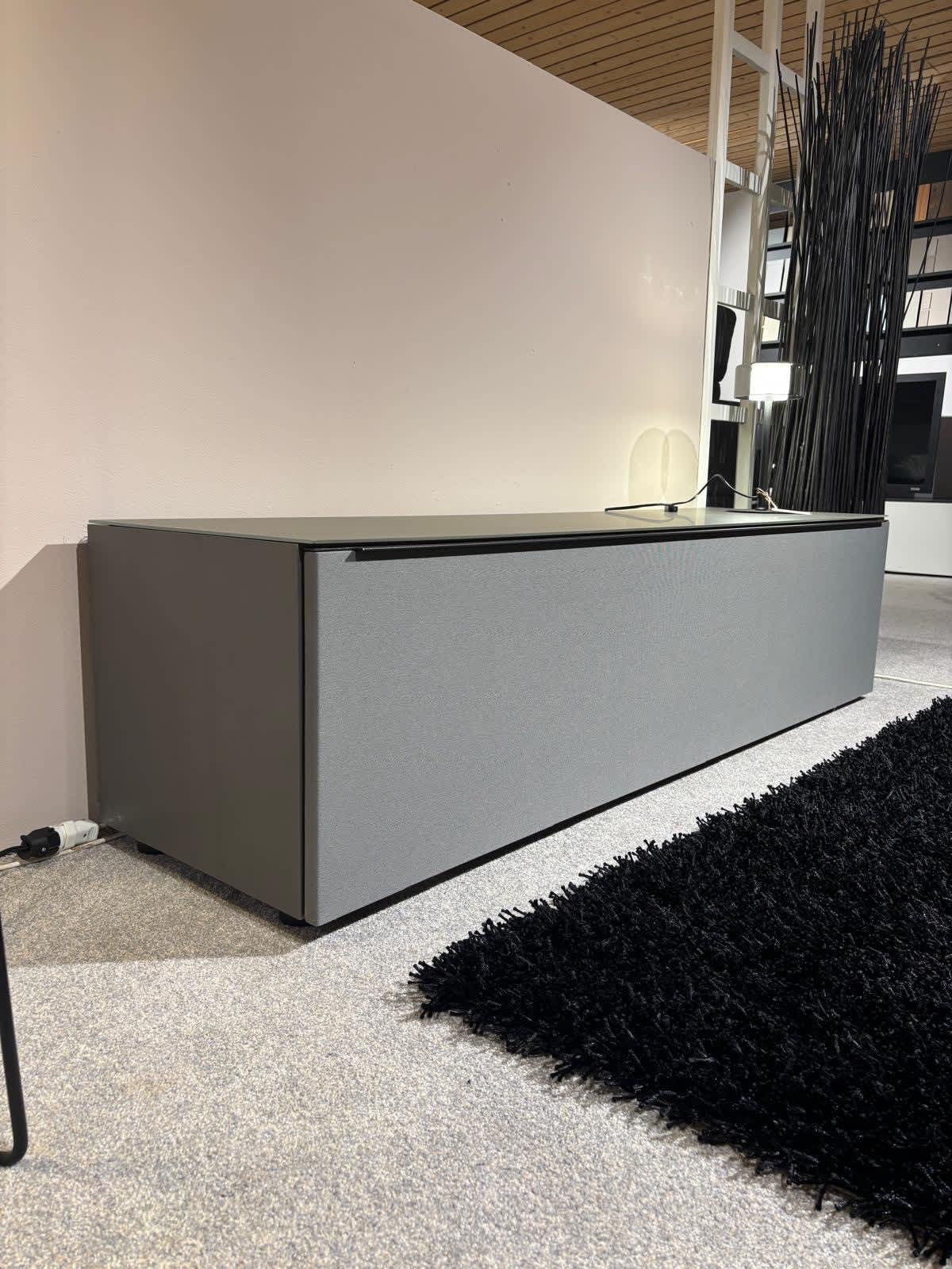Das Spectral Lowboard Bravo, ein moderner grauer Aufbewahrungsschrank, steht auf einem hellen Boden neben einem schwarzen Teppich in einem minimalistisch eingerichteten Raum. Das Spectral Lowboard Bravo, ein moderner grauer Aufbewahrungsschrank, steht auf einem hellen Boden neben einem schwarzen Teppich in einem minimalistisch eingerichteten Raum.
