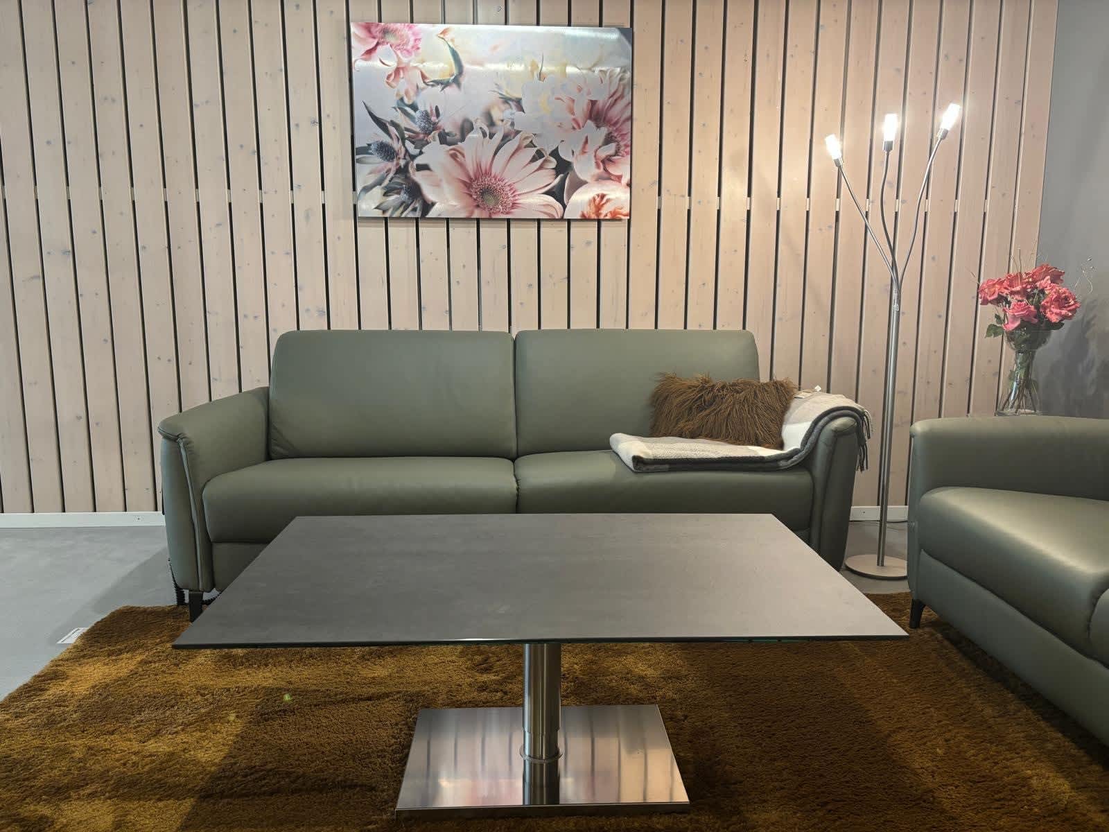 Ein modernes Wohnzimmer mit dem grünen Sofa „Ravenna“ aus der PM Oelsa Polstergruppe, einem schwarzen Tisch, einem Kunstwerk mit Blumenmotiv, einer Stehlampe und einem braunen Teppich. Ein modernes Wohnzimmer mit dem grünen Sofa „Ravenna“ aus der PM Oelsa Polstergruppe, einem schwarzen Tisch, einem Kunstwerk mit Blumenmotiv, einer Stehlampe und einem braunen Teppich.