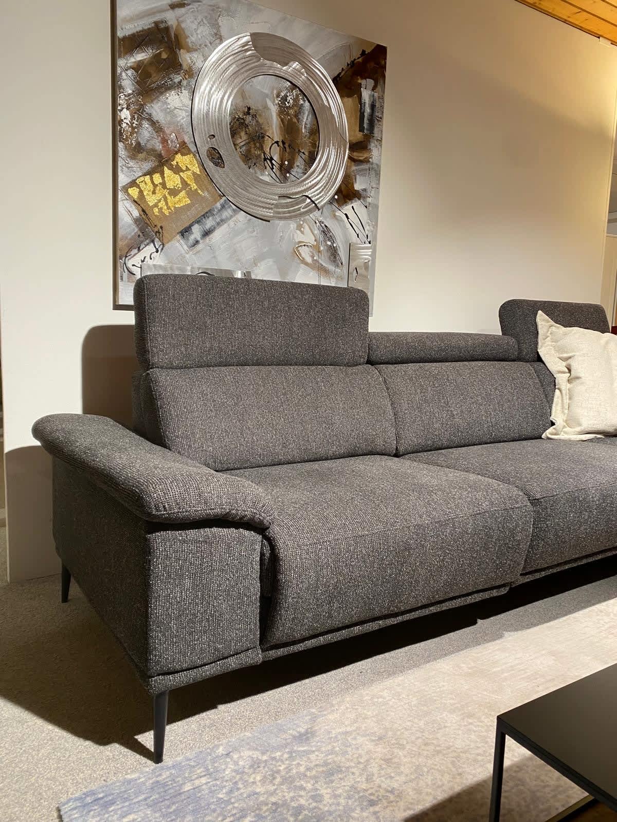 Das Dietsch-Sofa „Moon“ in modernem Grau mit einem hellen Kissen steht unter einem abstrakten Wandgemälde in einem hell beleuchteten Raum.