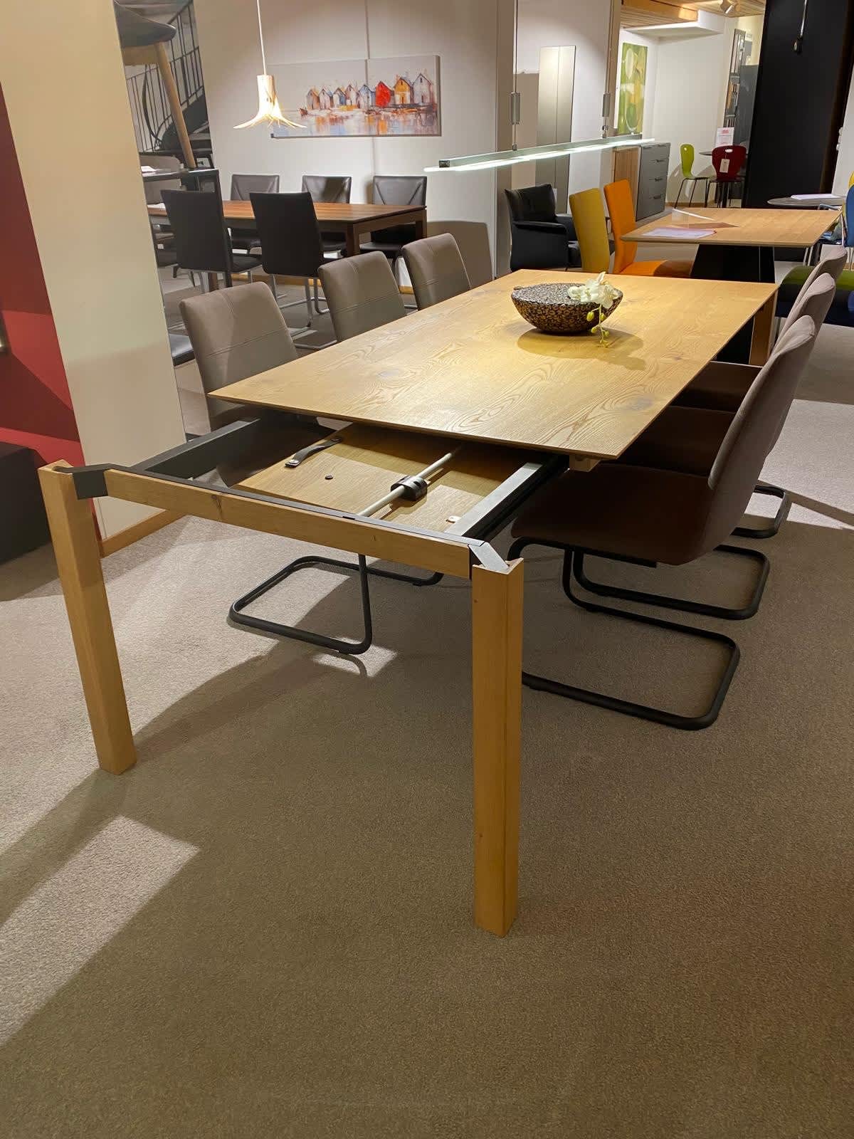 Der Venjakob-Auszugstisch Mod. 7, ein Esstisch aus Holz mit einer teilweise ausgezogenen Einlegeplatte, ist von modernen Stühlen umgeben. Der Venjakob-Auszugstisch Mod. 7, ein Esstisch aus Holz mit einer teilweise ausgezogenen Einlegeplatte, ist von modernen Stühlen umgeben.