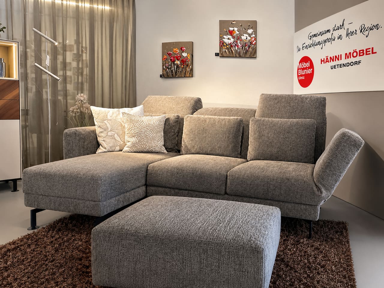Un moderne „Brühl Sofa Moule Small“ en gris avec des coussins se trouve sur un tapis brun, agrémenté de tableaux muraux et d’un panneau décoratif en arrière‑plan. Un moderne „Brühl Sofa Moule Small“ en gris avec des coussins se trouve sur un tapis brun, agrémenté de tableaux muraux et d’un panneau décoratif en arrière‑plan.