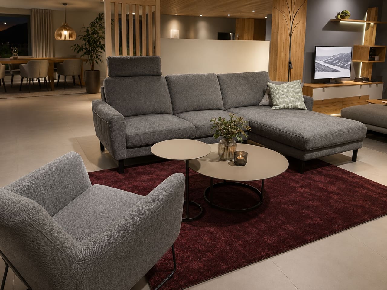 Modernes Wohnzimmer mit dem Sofa „Living“ in Grau aus der DFM-Polstergarnitur, einem Sessel, runden Couchtischen, einem roten Teppich und einem Fernseher auf einem Holzständer.