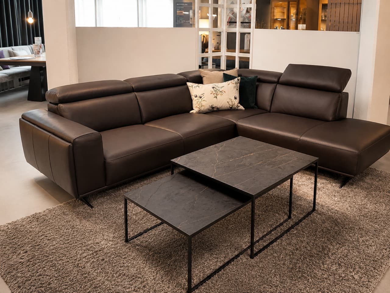 Ein modernes, braunes Leder-Ecksofa „Kent“ von Valmori mit Kissen, kombiniert mit zwei schwarzen Couchtischen und einem grauen Teppich im Wohnzimmer.