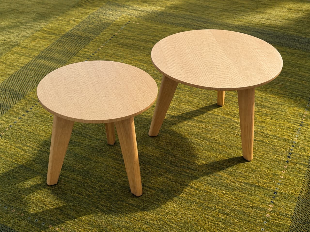 Deux tables basses rondes « Nordic » en bois, chacune avec trois pieds, sont posées sur un tapis à motif vert à la lumière du soleil. Deux tables basses rondes « Nordic » en bois, chacune avec trois pieds, sont posées sur un tapis à motif vert à la lumière du soleil.