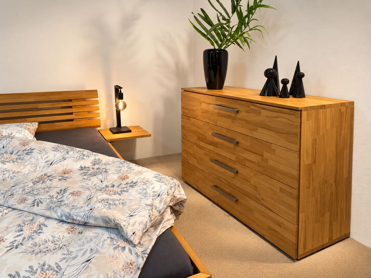 Eine Kommode „Covaro“ von Hasena aus Holz mit einer schwarzen Vase und Dekor sowie ein Bett mit geblümter Bettwäsche in einem modernen Schlafzimmer. Eine Kommode „Covaro“ von Hasena aus Holz mit einer schwarzen Vase und Dekor sowie ein Bett mit geblümter Bettwäsche in einem modernen Schlafzimmer.