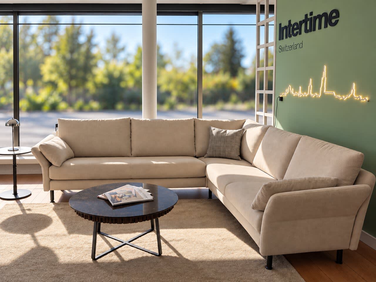 Ein Ecksofa „Campus“ in Beige von Intertime und ein runder Couchtisch in einem Raum mit großen Fenstern. Ein Ecksofa „Campus“ in Beige von Intertime und ein runder Couchtisch in einem Raum mit großen Fenstern.