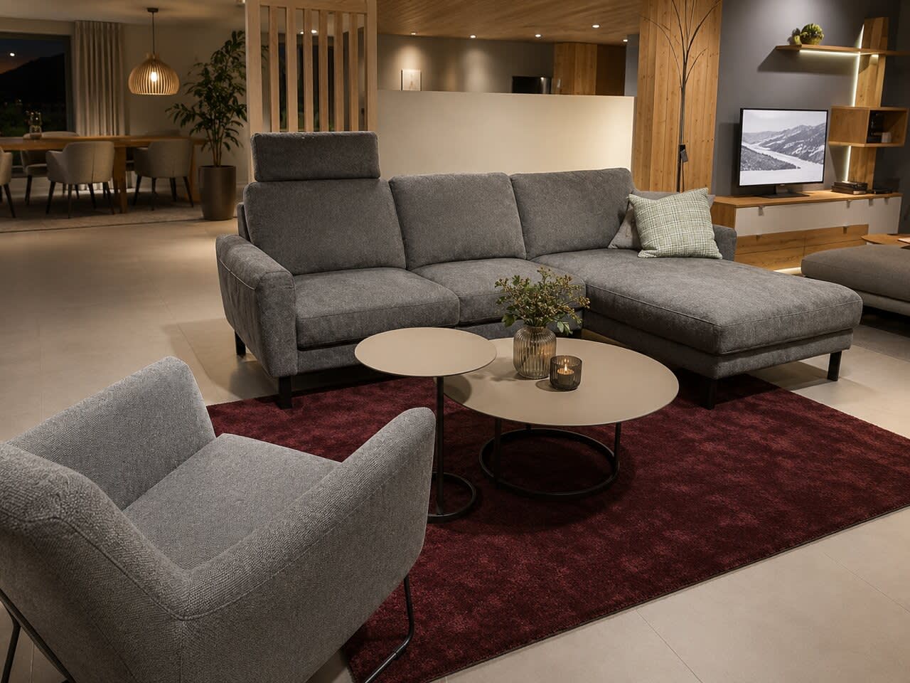 Modernes Wohnzimmer mit dem Sofa „Living“ in Grau aus der DFM-Polstergarnitur, einem Sessel, runden Couchtischen, einem roten Teppich und einem Fernseher auf einem Holzständer.