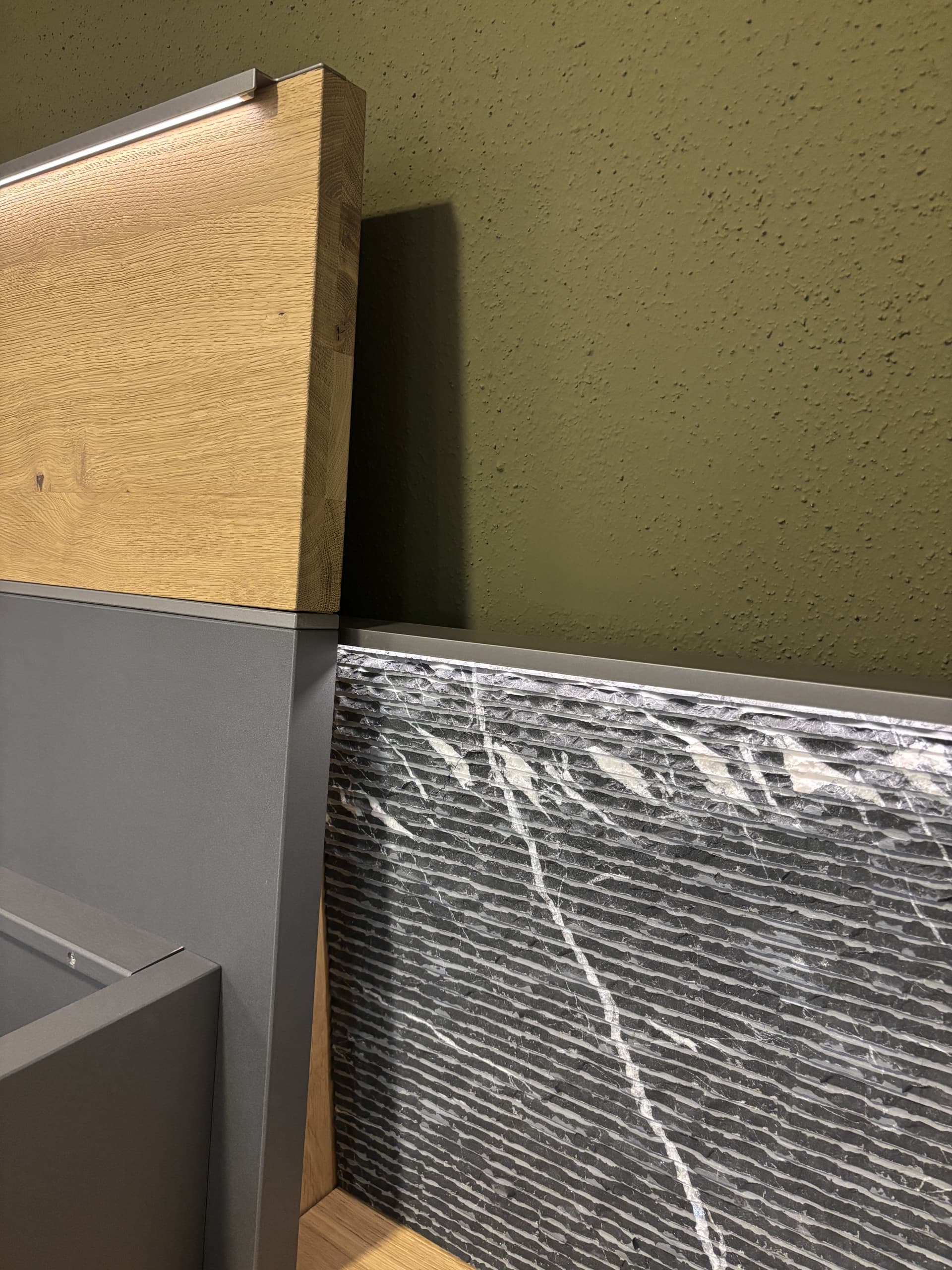 Modernes Innendesign mit dem Modell „Milo“ von Thielemeyer Schlafzimmer, gekennzeichnet durch Holzvertäfelungen, graue Steinplatten mit weißen Adern und eine olivgrüne Wand mit Struktur.