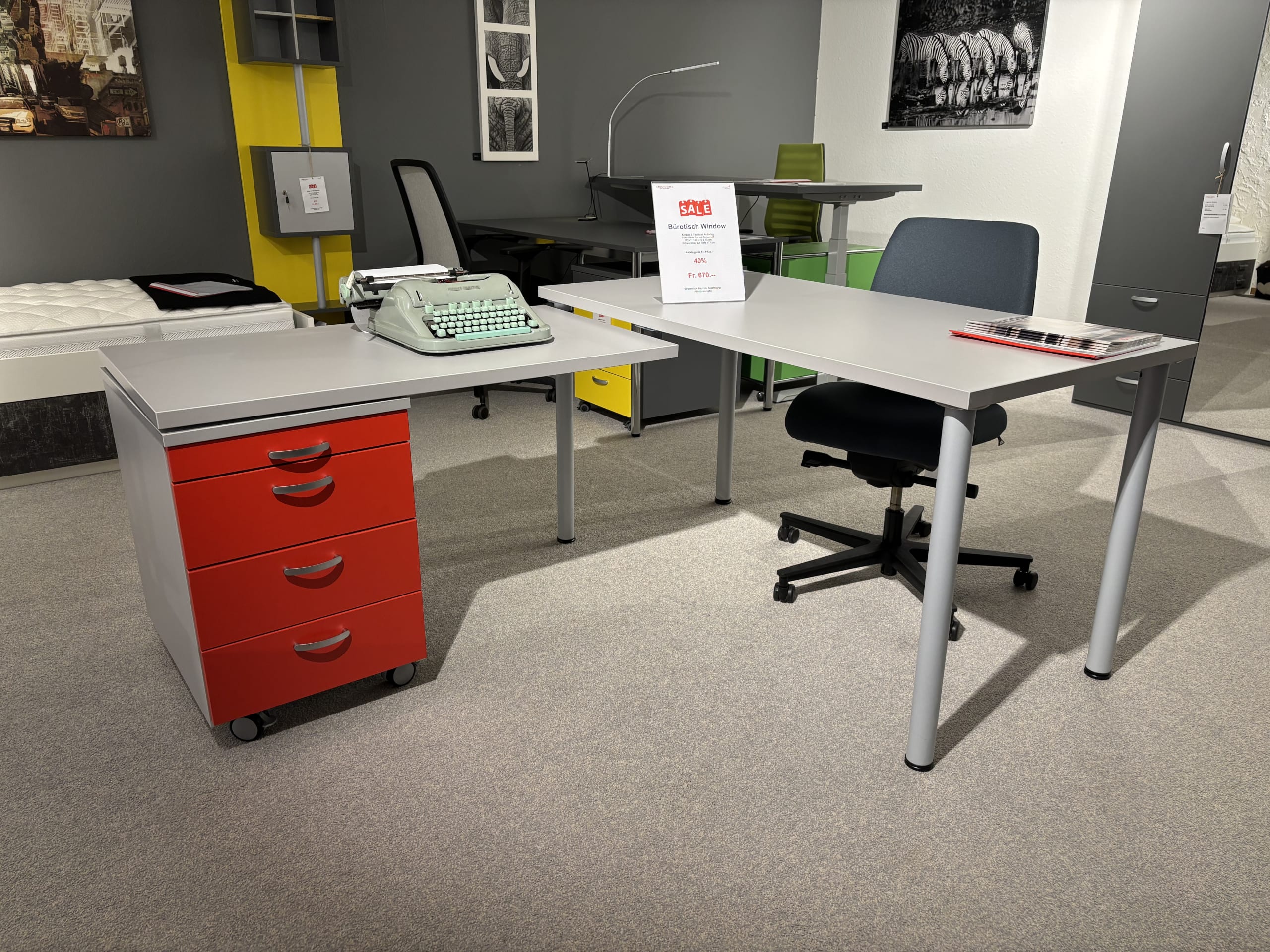 Mobilier de bureau composé de meubles de bureau avec 3 jardinières, un bureau gris, un caisson à tiroirs rouge, une machine à écrire, une chaise de bureau et un élément de décoration en arrière-plan.