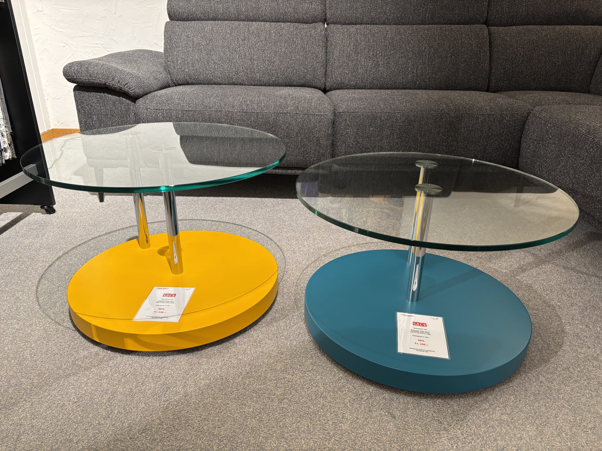 Zwei Beistelltische mit Glasplatten und gelb-blauen Untergestellen stehen auf einem grauen Teppich vor einem grauen Sofa. Produkt: Büromöbel mit 3 Boxen Window.