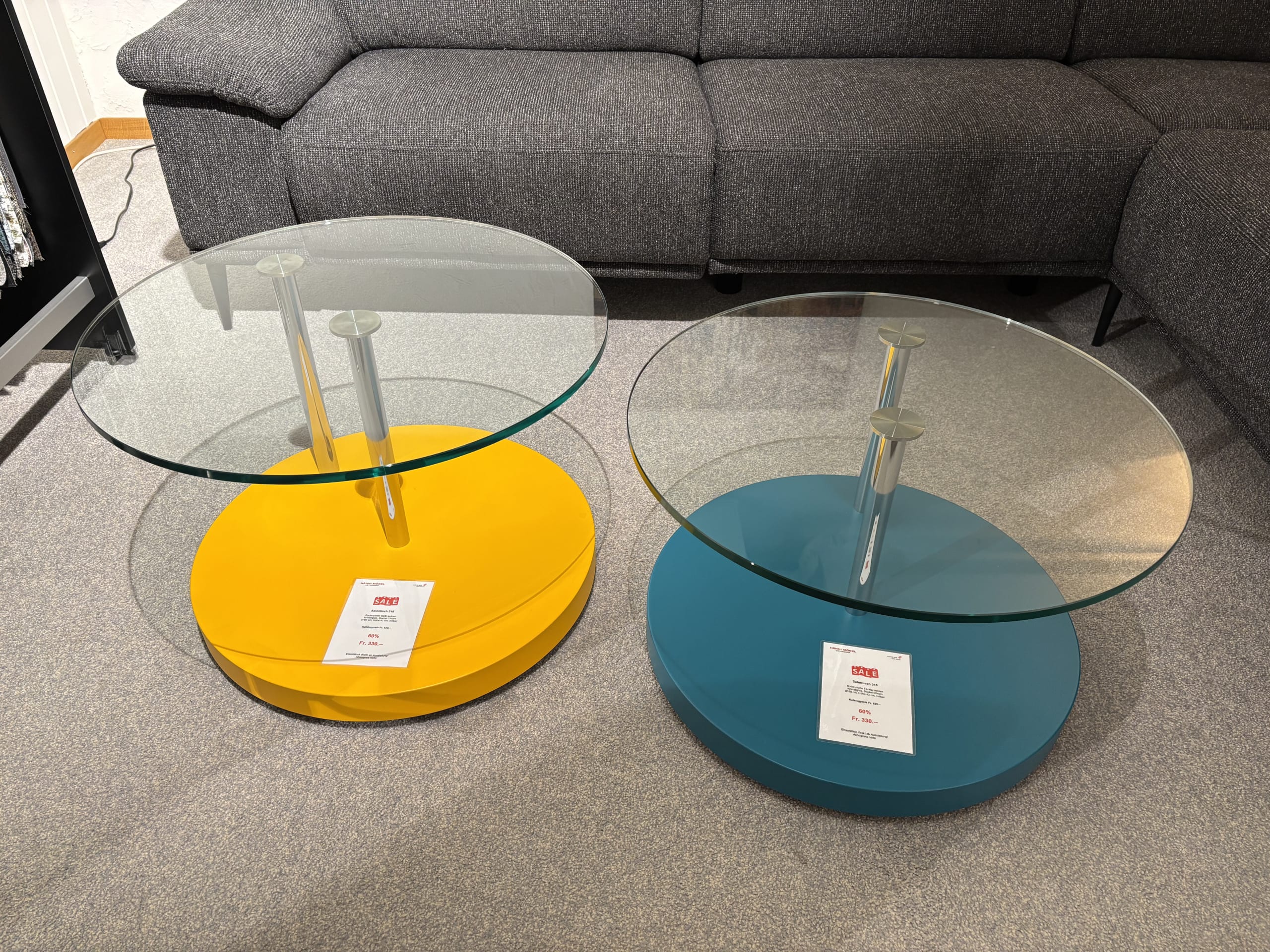 Zwei runde Couchtische aus Glas mit bunten Untergestellen, einer gelb und einer blau, vor einem grauen Sofa, ergänzen die Büromöbel mit 3 Boxen Window. Zwei runde Couchtische aus Glas mit bunten Untergestellen, einer gelb und einer blau, vor einem grauen Sofa, ergänzen die Büromöbel mit 3 Boxen Window.