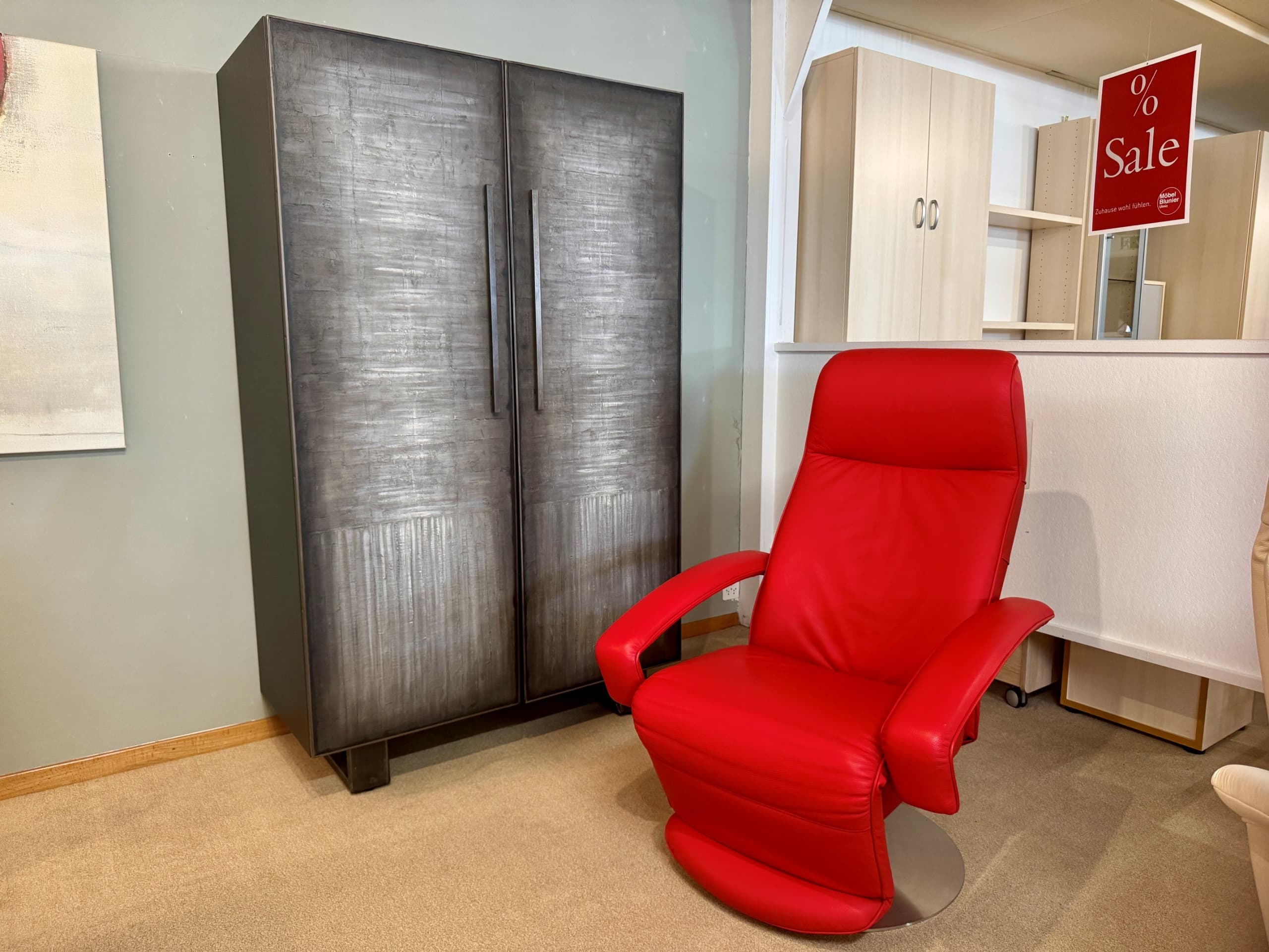 Neben dem „Sprenger Schrank 2-türig“, einem hohen, dunklen Schrank, steht ein roter Ledersessel in einem Ausstellungsraum, im Hintergrund ist ein rotes Verkaufsschild zu sehen.