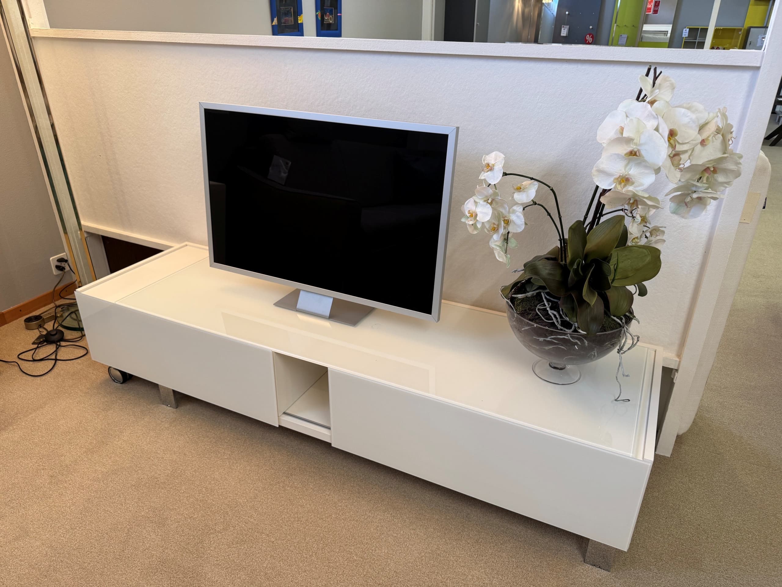Lowboard und Wandhängeschrank „D’Coll Wall“ mit einem Flachbildfernseher und einem Arrangement aus weißen Orchideen, platziert auf einem beigen Teppichboden.