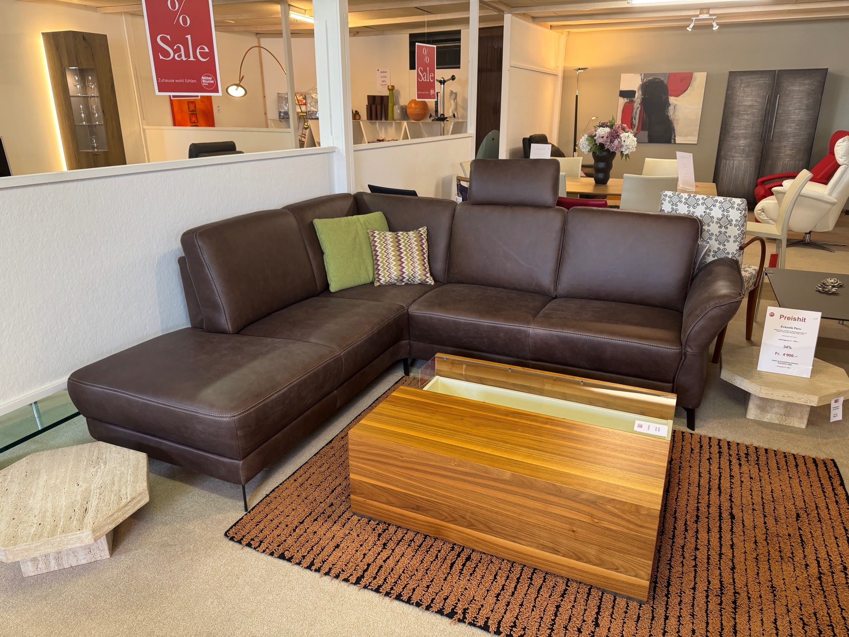 Das Ponsel Sofa Peru, eine braune Ledercouch mit bunten Kissen, steht neben einem hölzernen Couchtisch und Verkaufsschildern in einem Möbelhaus.