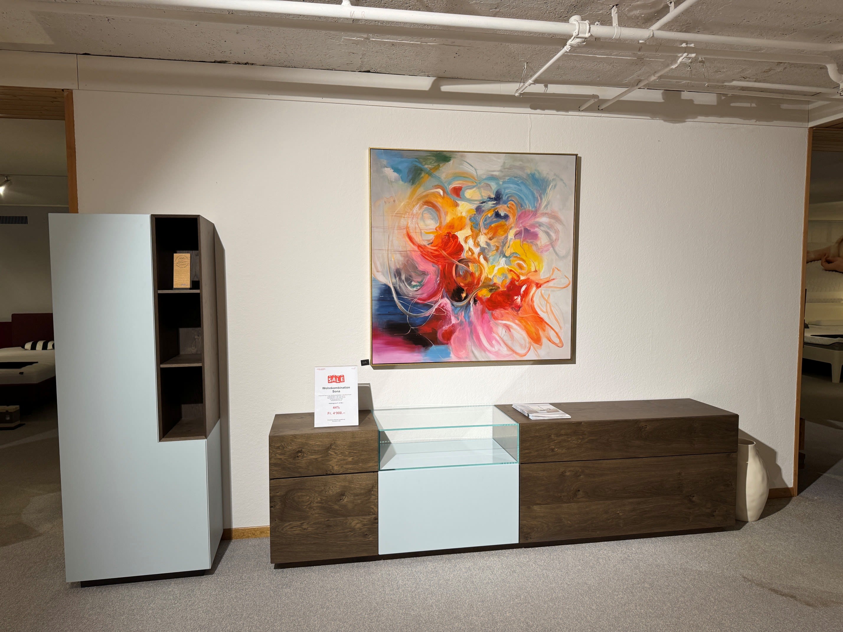 Die MAB Wohnkombination Sona mit Regal und Glasvitrine wird in einem Ausstellungsraum mit einem abstrakten, farbenfrohen Kunstwerk darüber präsentiert. Die MAB Wohnkombination Sona mit Regal und Glasvitrine wird in einem Ausstellungsraum mit einem abstrakten, farbenfrohen Kunstwerk darüber präsentiert.