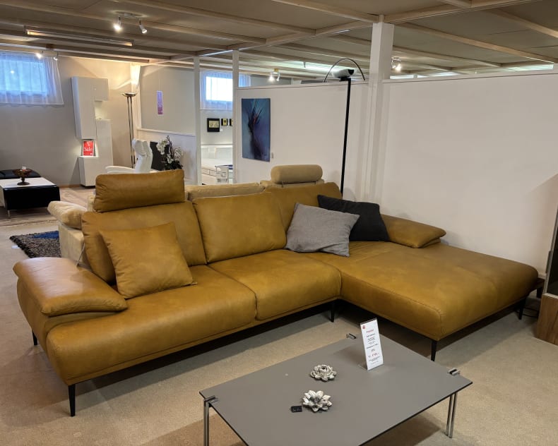 Die Polstergruppe Samiraa, ein senfgelbes Sofa mit grauen Kissen, wird in einem Ausstellungsraum präsentiert.