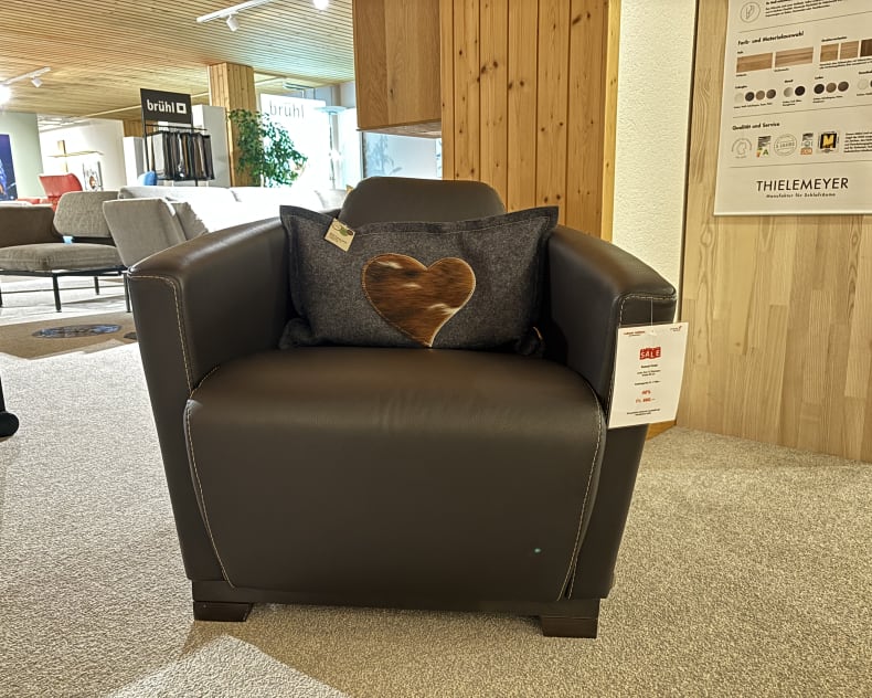 Ein Büromöbel mit 3 Boxen Window, bestehend aus einem dunklen Ledersessel mit einem Kissen mit Herzmuster, wird in einem modernen Ausstellungsraum ausgestellt.