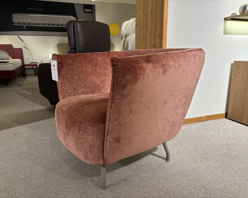 Auf einem grauen Teppich in einem Ausstellungsraum mit Betten und einem braunen Stuhl sorgt der rosa Samtstuhl neben den Büromöbeln mit 3 Boxen Window für Eleganz.