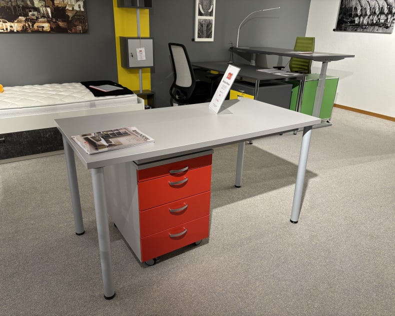 Showroom contemporain avec mobilier de bureau comprenant trois vitrines, un bureau gris, des tiroirs rouges, une chaise et des étagères colorées. De la moquette et un matelas à proximité complètent l'ambiance.