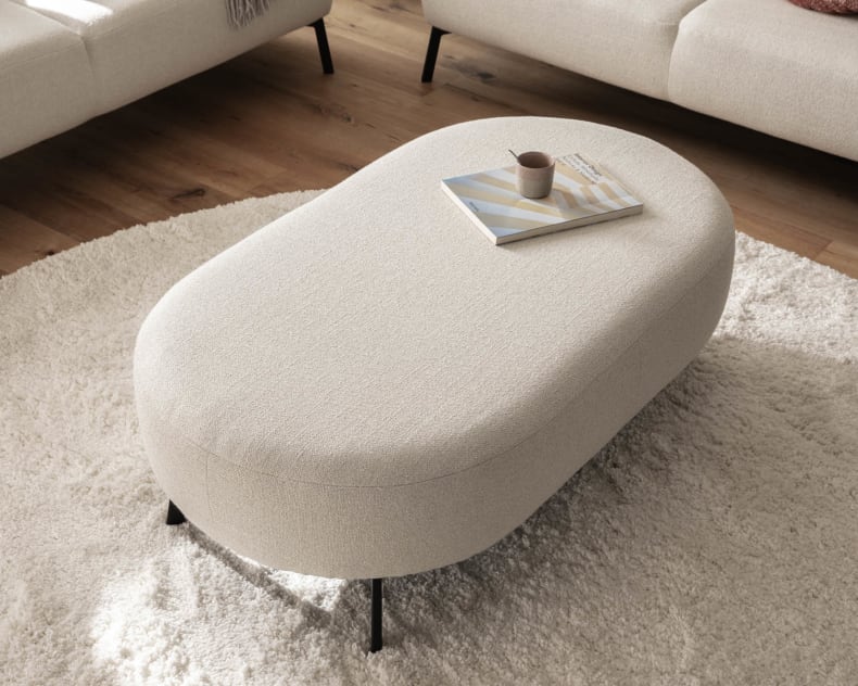 Das Candy Sofa Nimba dient als perfekte Kulisse für einen ovalen beigen Ottomanen, auf dem ein Buch und eine Tasse liegen, die alle auf einem runden beigen Teppich im Wohnzimmer ruhen.