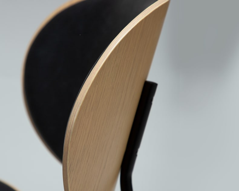 Gros plan sur une chaise Clementina de Tisch Ooid & Stühle, avec un dossier en bois incurvé et une assise rembourrée noire, sur fond clair.
