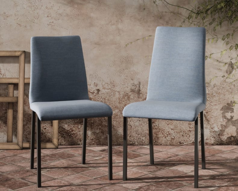 Deux chaises Willisau Leni, recouvertes de tissu bleu et dotées de pieds noirs, sont posées sur un sol carrelé devant un mur rustique orné de vignes.