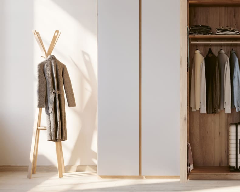 Le portant à vêtements Wimmer se dresse au soleil à côté d'une armoire minimaliste avec des compartiments ouverts et fermés où sont suspendus des vêtements, tandis qu'un manteau est drapé sur le support en bois.