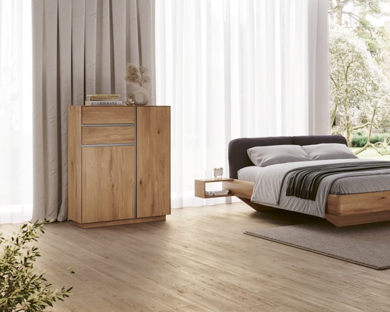 Minimalistisches Schlafzimmer mit einem Holzbett, einem Nachttisch, großen Fenstern mit hellen Vorhängen und einem Wimmer Highboard-Schrank.