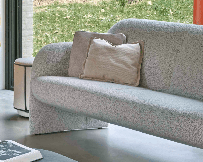 Leolux Sofa Modius in Hellgrau mit zwei beigen Kissen, positioniert an einem Fenster mit Blick auf eine grüne Wiese.