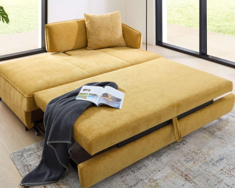 Das Bali Bettsofa Ilka, ein gelbes ausziehbares Schlafsofa mit Kissen, Decke und einer aufgeschlagenen Zeitschrift, steht an großen Fenstern.