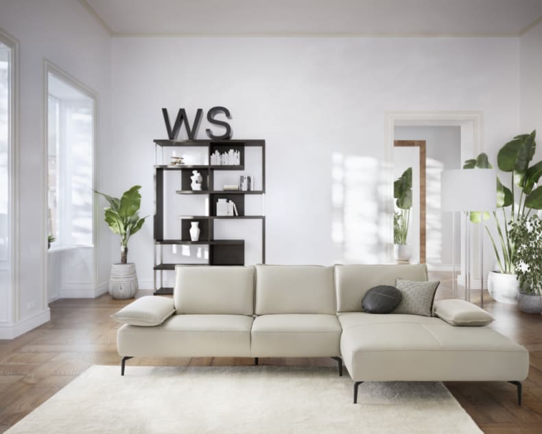 Modernes Wohnzimmer mit einem W.Schillig Sofa Caipirinjaa 16568 in Beige, großen Fenstern, Grünpflanzen und einem schwarzen Regal mit "WS"-Dekor.