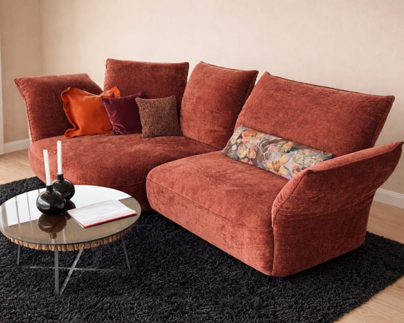 Das rote Samtsofa der Koinor Polstergruppe Abby mit verschiedenen Kissen steht auf einem schwarzen Teppich neben einem runden Couchtisch aus Glas, auf dem Kerzen und ein Buch stehen.