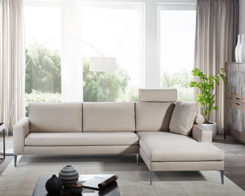 Modernes Wohnzimmer mit einem Artanova Sofa Castell II 9132, großen Fenstern, neutralem Dekor und Zimmerpflanzen.