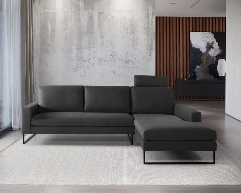 Modernes Wohnzimmer mit dem Artanova Sofa Castell II 9132 in Dunkelgrau auf einem hellen Teppich, unterstrichen durch abstrakte Wandkunst und raumhohe Fenster.