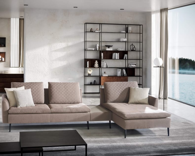 Modernes Wohnzimmer mit dem Artanova Sofa Dyo 9192, Regalen, großen Fenstern und einem malerischen Blick auf den See.