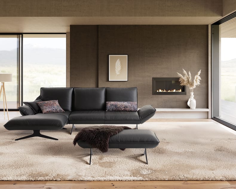 Modernes Wohnzimmer mit dem Koinor Sofa Buddy in Schwarz, kombiniert mit einer Ottomane, einem Kamin, einer Stehlampe und großen Fenstern, die einen Ausblick auf die Landschaft bieten.