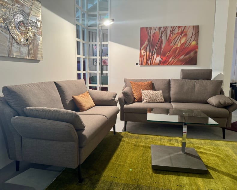 Modernes Wohnzimmer mit zwei Horst-Sofas Evita 3303 in Grau, einem Couchtisch aus Glas, einem grünen Teppich und abstrakter Wandkunst.