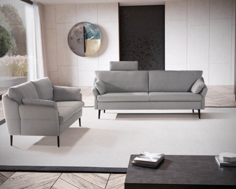 Modernes Wohnzimmer mit zwei Horst-Sofas Evita 3303 in Grau, einem hellen Teppich, Wandkunst, Bücherregalen und einem großen Fenster mit transparenten Vorhängen.