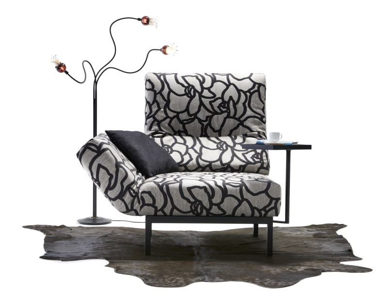 Das Signet Sofa Chimba zeichnet sich durch ein modernes Design mit einem auffälligen Blumenmuster aus, gepaart mit einem schwarzen Kissen, einem Beistelltisch, einer Stehlampe und einem Kuhfellteppich auf weißem Hintergrund.