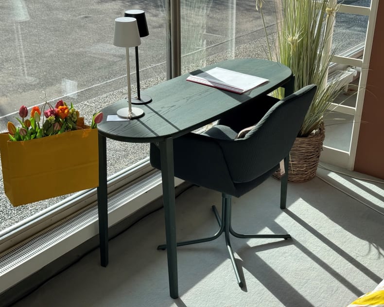 Der Mobitec Tisch Dot T0400, zusammen mit einem Stuhl am Fenster, zeigt eine Lampe, ein Notizbuch und einen Stift, umgeben von Pflanzen in der Nähe.