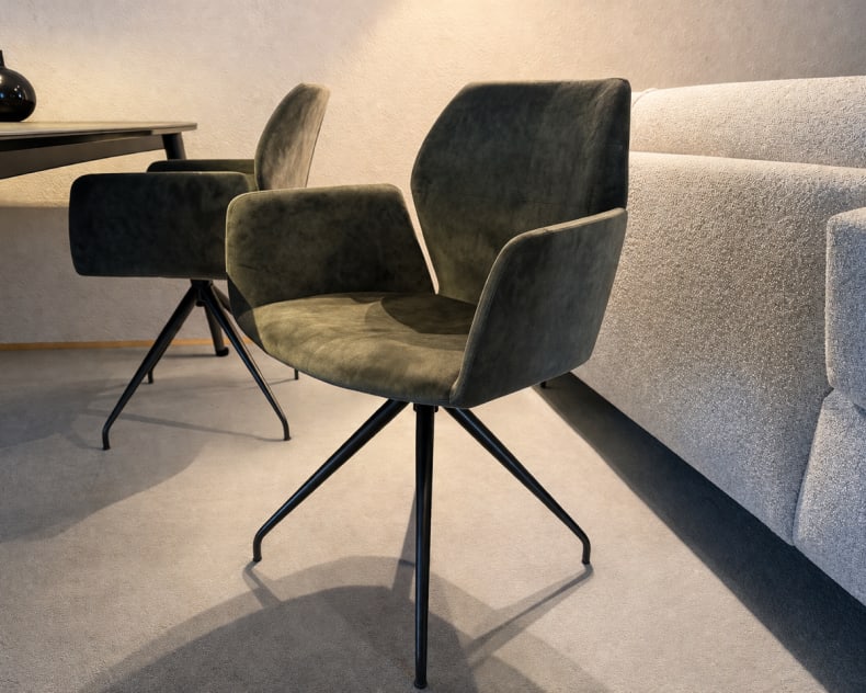 Der Mobitec Stuhl Mood 95, ein moderner grüner Drehstuhl mit Metallbeinen, steht neben einem grauen Sofa und einem dunklen Tisch in einem modernen Raum.