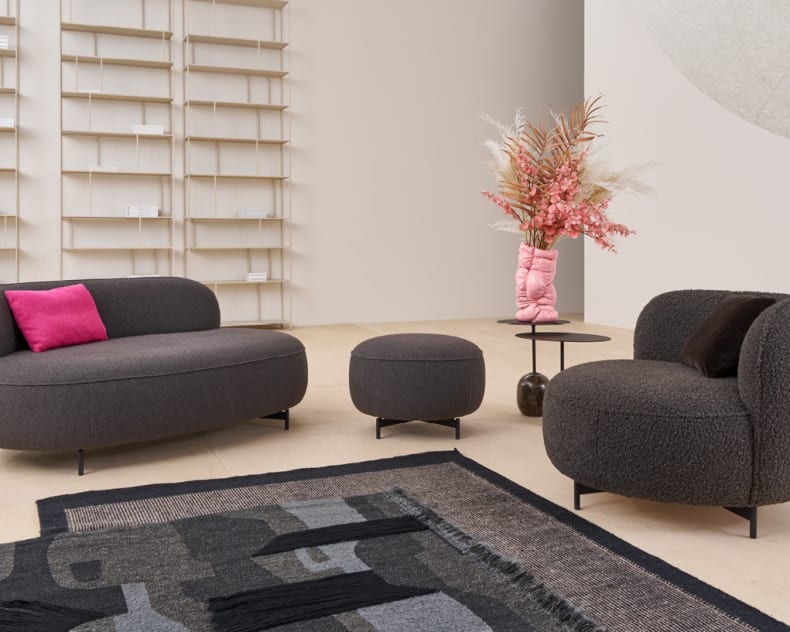 Modernes Wohnzimmer mit dem Himolla Einzelsessel 7596, dunkelgrauem Sofa, Ottomane, rosa Kissen und einer Blumenvase auf einem runden Tisch.