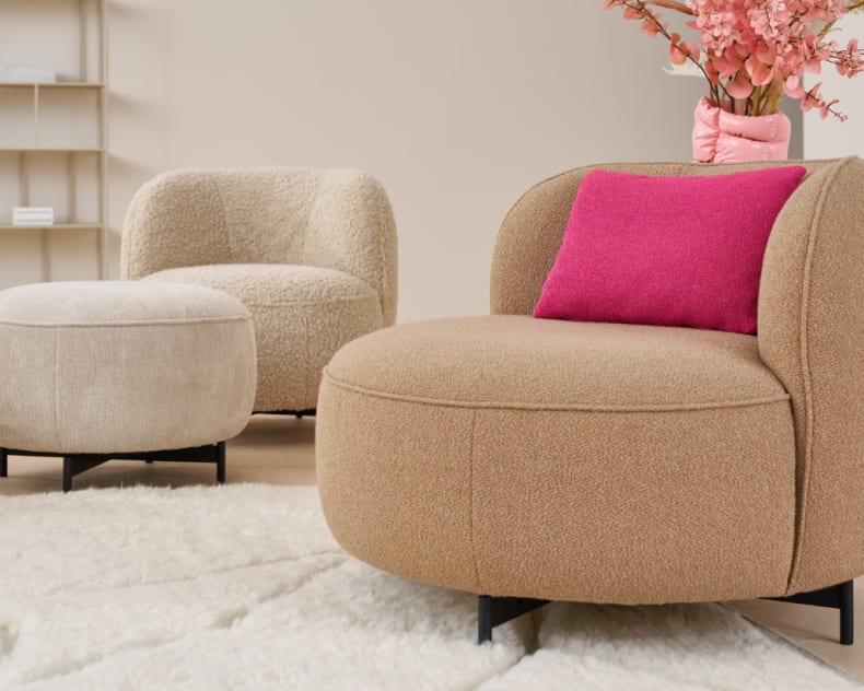 Der Himolla Einzelsessel 7596 ist ein moderner beiger Sessel mit einem rosa Kissen, einem passenden Ottomanen, einem Teppich und Blumen in einer Vase, alles auf einem hellen Boden.