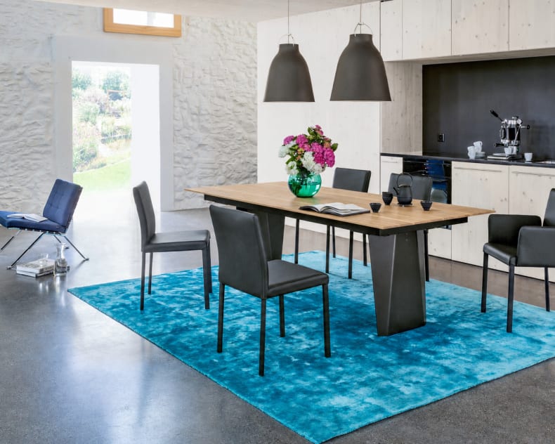Moderne Essecke mit sechs schwarzen Stühlen von Girsberger Stuhl Kuba, einem Holztisch, Pendelleuchten, einem blauen Teppich und einer Blumenvase.