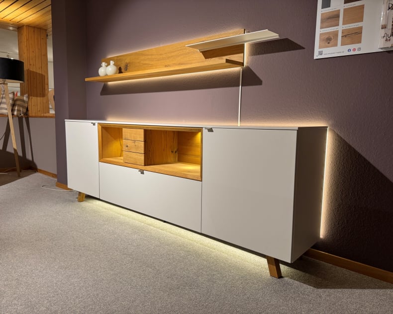 Das Venjakob Wohnprogramm Andiamo Home Sideboard besticht durch sein modernes weißes Design mit Holzakzenten, LED-Beleuchtung und einem an der Wand befestigten Regal, das jeden Raum stilvoll aufwertet.